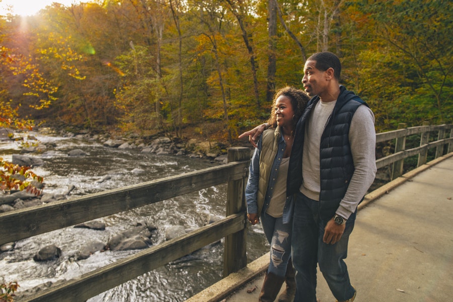 A couple walking on a bridge over a scenic river with colorful autumn foliage.