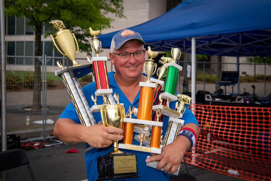 BBQ Contest Winner: A man smiling broadly, holding several colorful trophies, including a prominent gold trophy, at the BBQ battle competition.