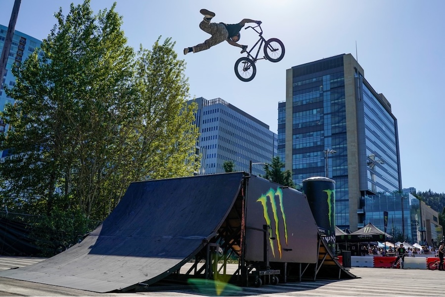 BMX Stunt Show: A BMX rider performing a mid-air stunt over a ramp in an urban setting, with high-rise buildings and a blue sky in the background.