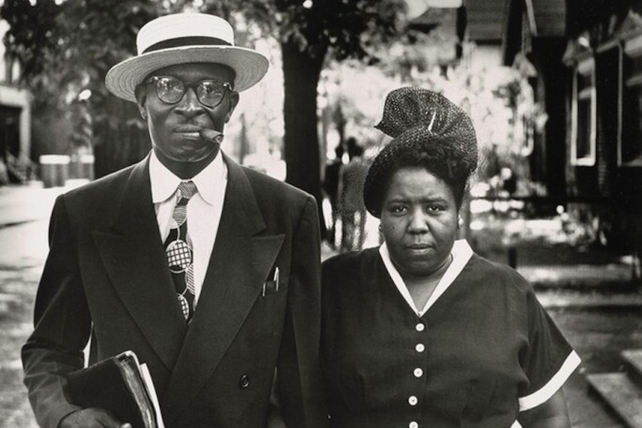 A black and white photo of a couple. The man is wearing a suit and hat, smoking a cigar, and holding a book. The woman stands next to him, wearing a dress and looking directly at the camera.