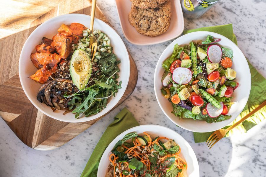 This image features three colorful bowls filled with fresh salads and vegetables, along with a plate of cookies and a can of drink, all set on a marble table.