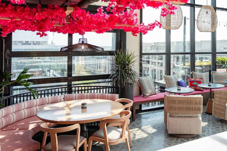 Indoor seating area with large windows and vibrant red flowers on the ceiling. Features round tables, cushioned chairs, a bench with pillows, and wicker pendant lights.