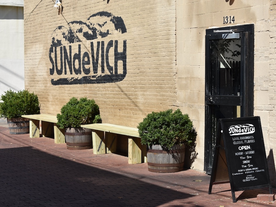 The exterior of SUNdeVICH, a sandwich shop with a tan brick wall featuring the restaurant's logo painted in black. Wooden benches with potted plants are placed along the wall. A black chalkboard sign near the entrance displays the shop's hours of operation.