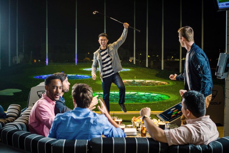 Group of friends enjoying a night at Topgolf, with one person celebrating a successful swing while others watch and cheer from their lounge area.