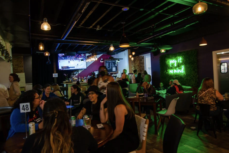 A lively bar scene with patrons sitting at tables, chatting, and enjoying drinks. The bar area is illuminated by hanging lights, with a neon sign on the wall reading "pay it no mind." A television in the background displays a sports game, adding to the energetic atmosphere.