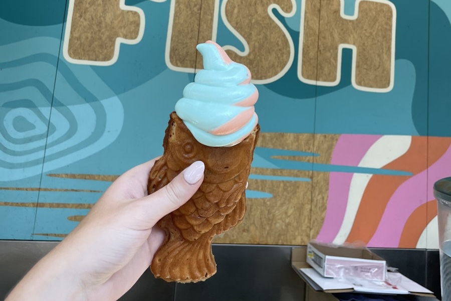 A hand holds a fish-shaped waffle cone filled with swirled pastel blue and pink soft-serve ice cream, in front of a colorful background with the word 'FISH' partially visible.