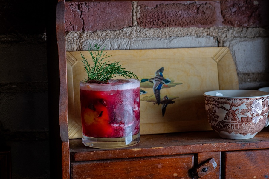 A vibrant red cocktail garnished with a sprig of greenery sits on a rustic wooden shelf, next to a decorative ceramic cup and a painted illustration of mallard ducks in flight.