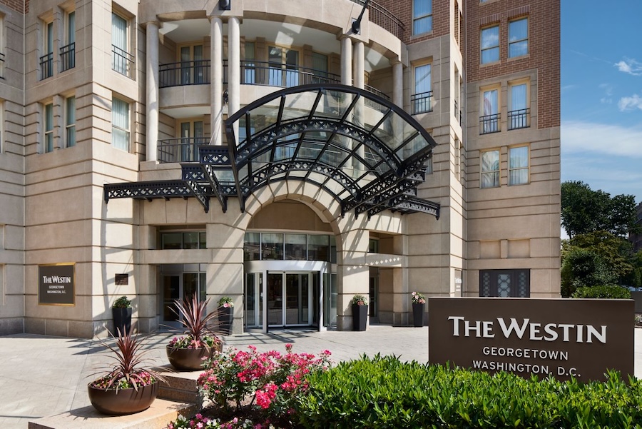 The grand entrance of The Westin Georgetown, Washington, D.C., showcasing a stylish glass canopy, a lush floral arrangement, and the hotel's name prominently displayed on a sign.