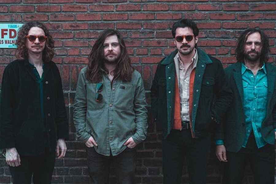 A four-member band poses in front of a brick wall, each dressed in casual jackets and shirts, with a laid-back, retro vibe. Two of the members wear sunglasses, and their long hair and beards contribute to a rock or indie style.