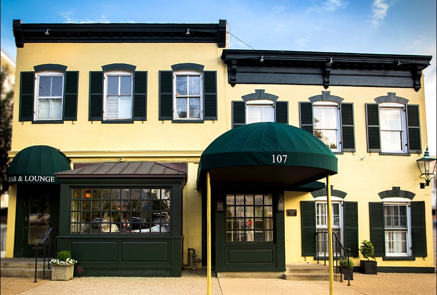 The exterior of a charming yellow building features dark green accents, including shutters and an awning, with large windows and a welcoming entrance labeled "Bar & Lounge." The building is set against a clear blue sky, showcasing its inviting atmosphere.