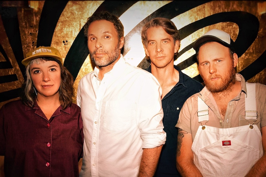 A four-member band poses in front of a stylized background with golden tones and black swirls. The group has a casual look, with one member wearing a cap and another in overalls, creating an indie or alternative vibe.