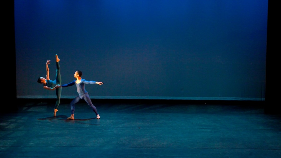 Two dancers perform an elegant contemporary ballet duet on a dimly lit stage, with one dancer balancing gracefully on one leg while the other supports them, both dressed in sleek blue costumes.