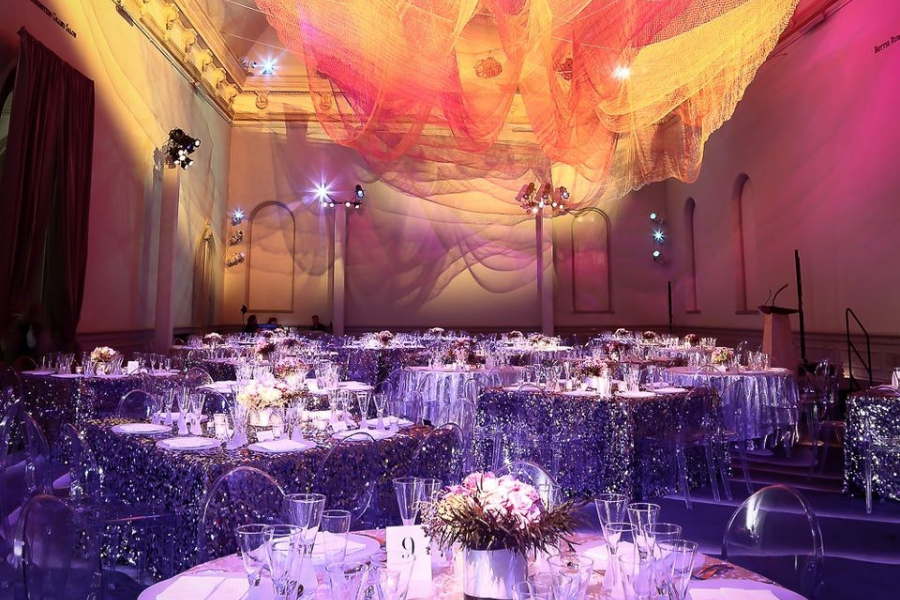 A grand event space with tables set for a formal dinner, featuring shimmering tablecloths and elegant glassware. Overhead, a colorful, flowing art installation made of fabric or netting is illuminated in shades of pink, orange, and yellow, creating a dramatic visual display against the high ceiling.