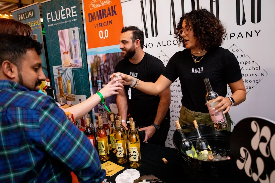 A lively event featuring a booth for non-alcoholic beverages, with two representatives serving samples of various bottled drinks to attendees. Colorful banners for brands like "Fluère" and "Damrak Virgin 0.0" are displayed in the background.