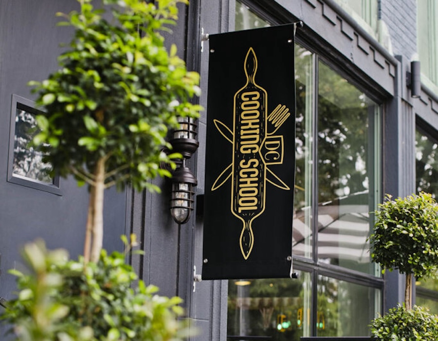 A black and gold sign for DC Cooking School hangs outside a modern building with greenery.