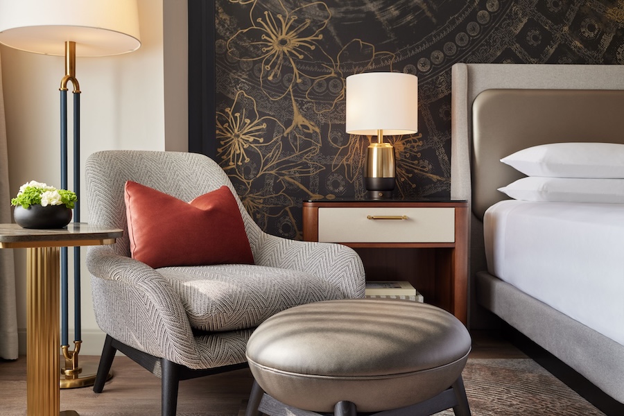 A cozy hotel room corner featuring a textured armchair, a gold-accented lamp and a plush bed with elegant decor.