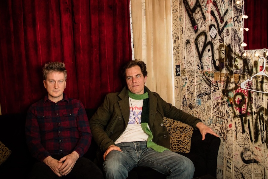 Two men sit on a black couch in a dimly lit room with red velvet curtains and graffiti-covered walls, exuding a casual yet intense presence.