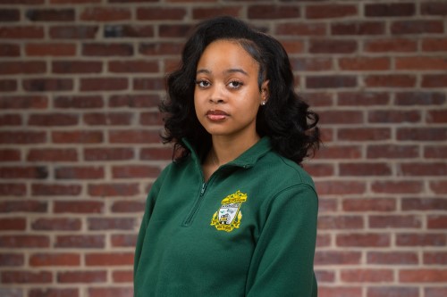 The professional headshot of Tabitha, a young Black woman. She is wearing a her Academy polo from Jackson-Reed High School and stands in front of a brick wall. 