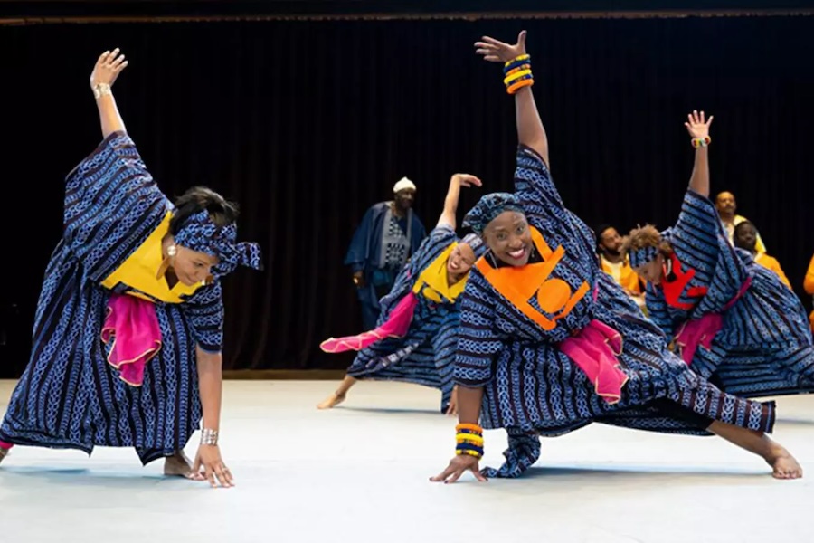 A group of dancers in vibrant blue and pink West African attire perform a high-energy routine on stage with drummers in the background.