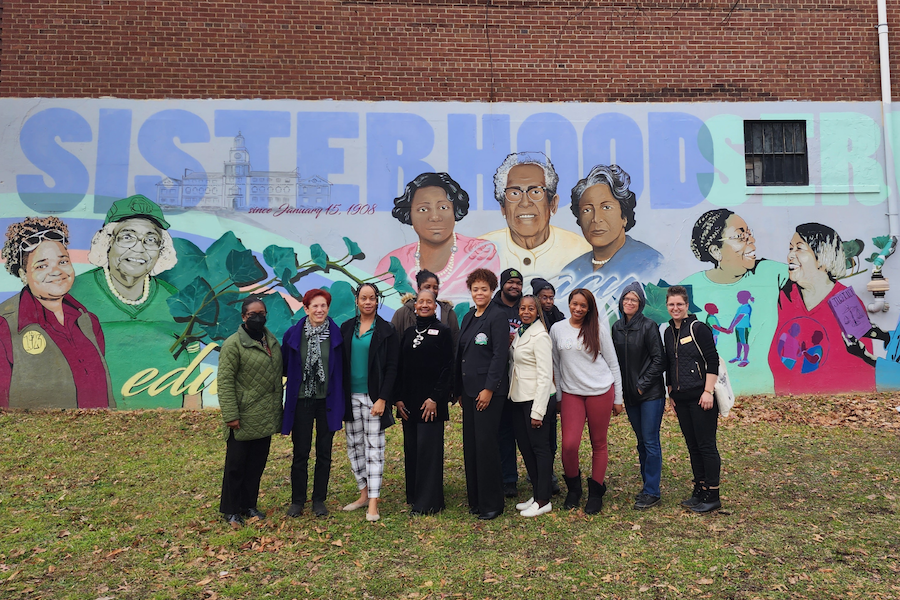 A group of women poses in front of a mural that says, "Sisterhood."