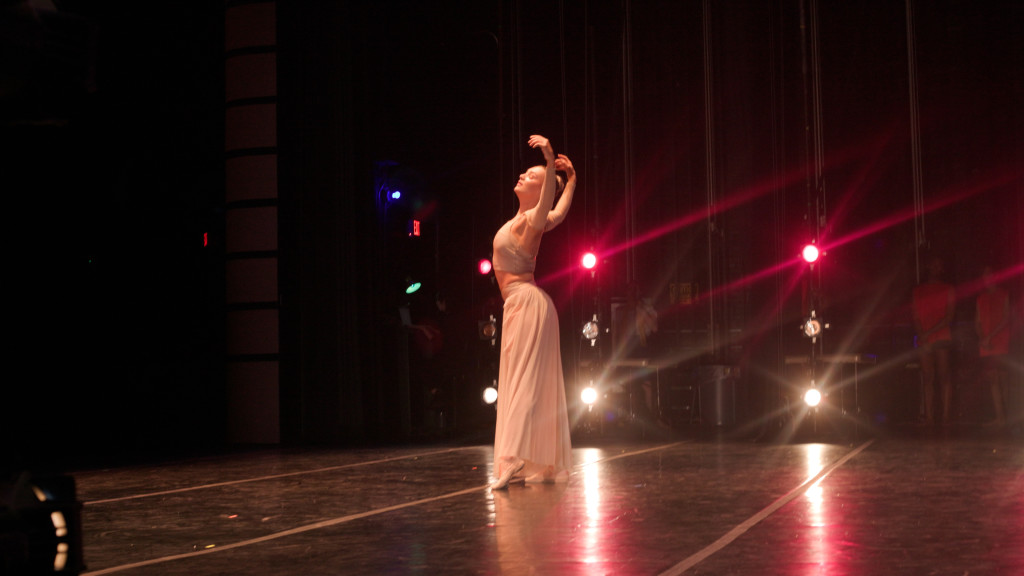 A dancer on stage, illuminated by spotlights, strikes a graceful pose with arms raised. She is wearing a flowing, light-colored outfit, and the stage is dark except for the bright lights behind her.