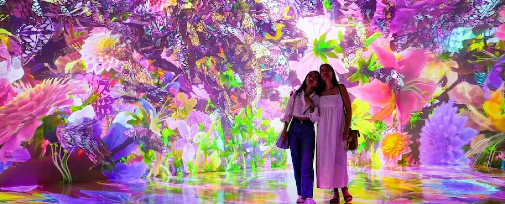 Two visitors stand in a vibrant, immersive digital art installation at ARTECHOUSE, surrounded by colorful, glowing flowers and butterflies.