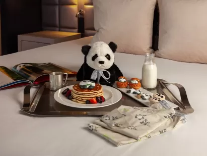 Stuffed animal panda sitting on a crisp, white hotel bed with panda-themed treats.