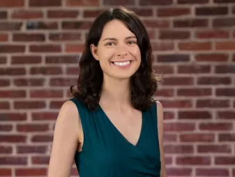 A woman with short brown hair, wearing a green sleeveless top, stands in front of a brick wall.